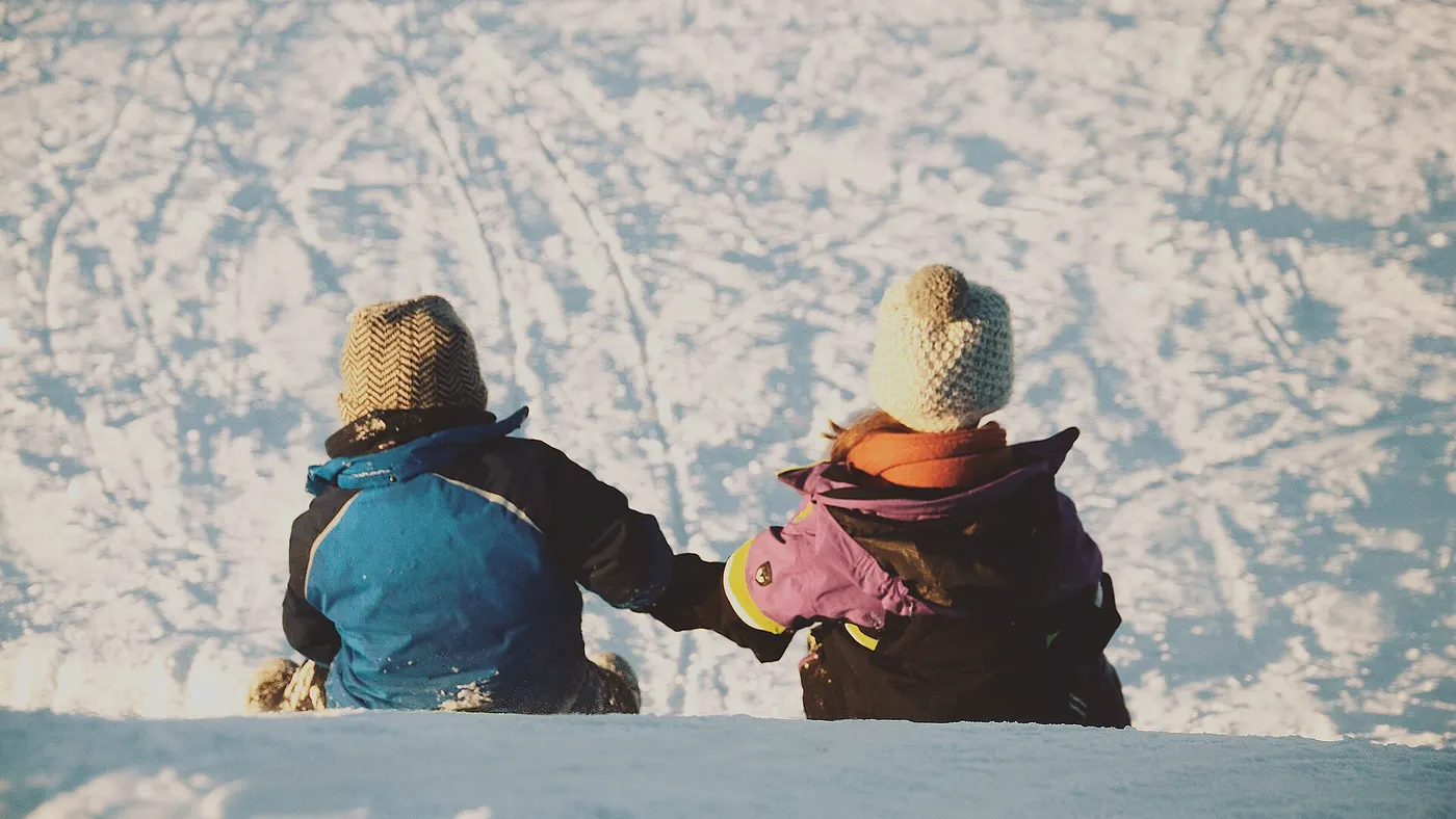 Deux enfants en bonnet assis dans la neige, vus de dos