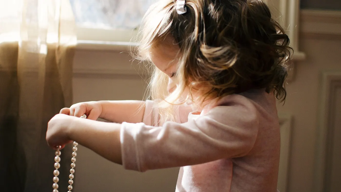 Une petite fille qui manipule un collier de perles près d’une fenêtre